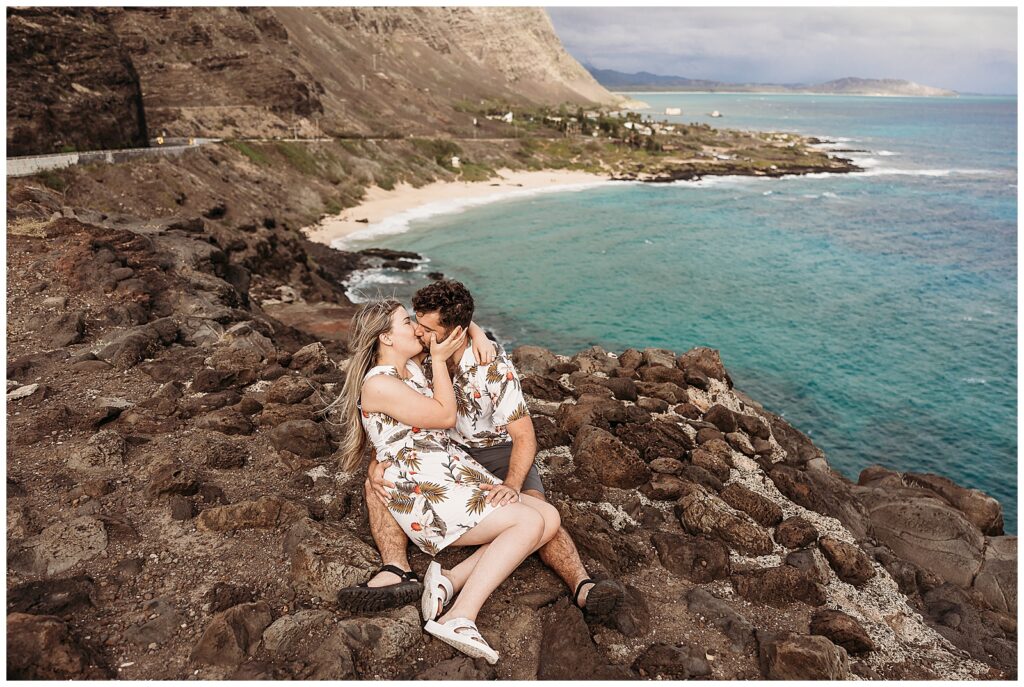 couple sitting on ledge of lava rocks kissing with makapuu beach in the background